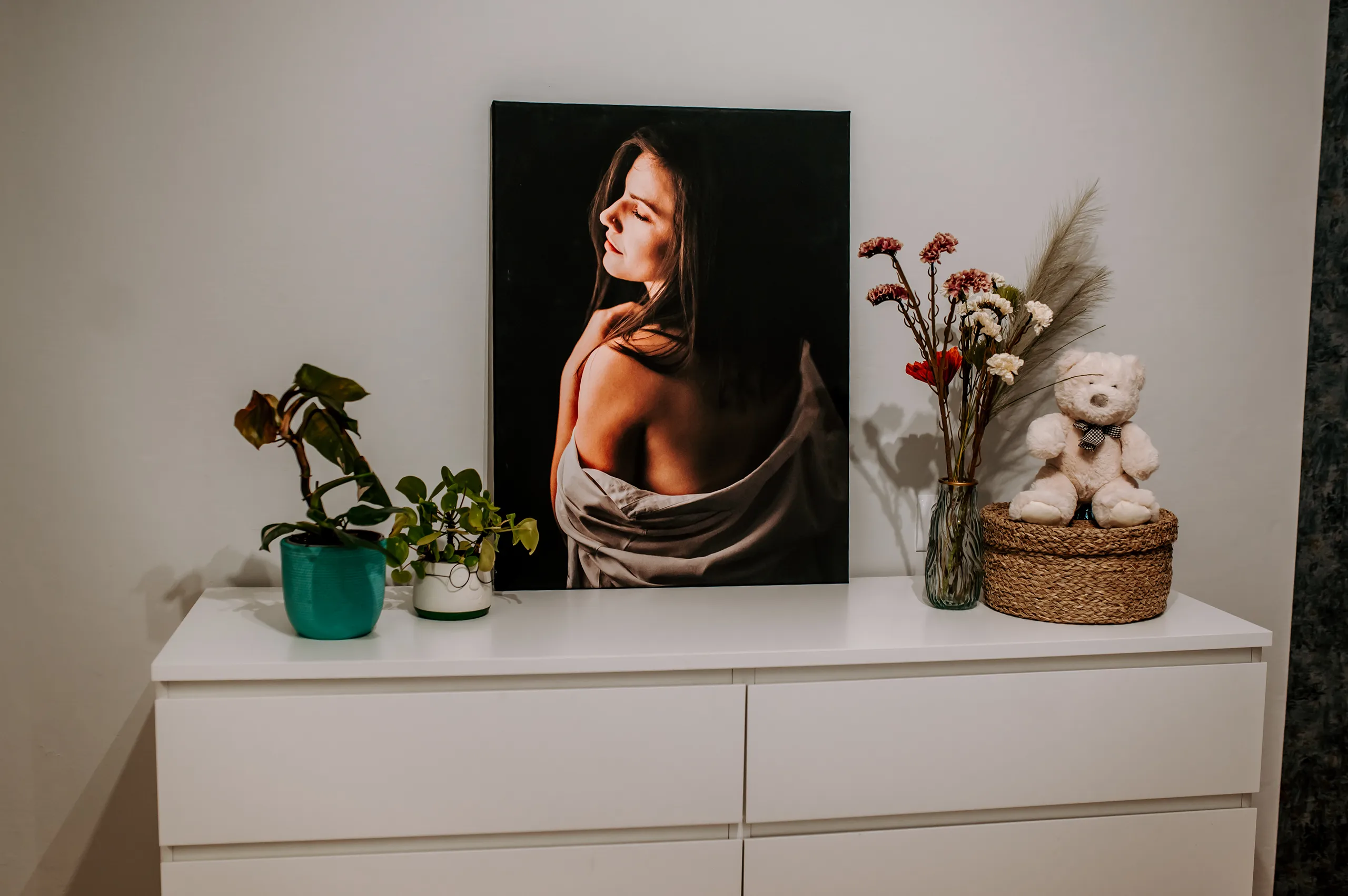 Fotoobraz z portretem kobiety z nagim ramieniem na czarnym tle ustawiony na białej komodzie